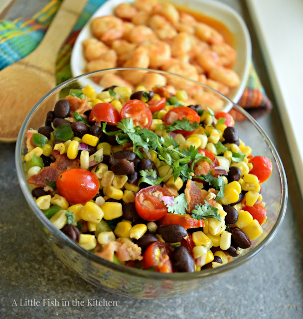 A richly-colored corn and black bean salad sits in a clear glass serving bowl. A green and blue plaid tea towel sits near to the bowl with a wooden spoon resting on top. A platter of grilled shrimp sits behind the bowl of corn salad, it's slightly blurred. The corn and black bean salad has brightly colored ingredients that include yellow corn, red ripe cherry tomatoes, green fresh peppers, bacon bits and hearty black beans. 
