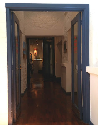 View through a blue framed doorway, down a polished wooden floorboard corridor with white walls. In the distance is another blue framed doorway in the centre of which one can see a light mannequin torso in the spotlight wearing a coral pink bikini.