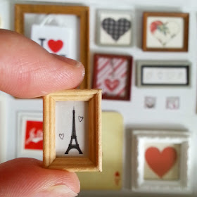 Modern miniature framed sketch of the Eiffel tower being held between thumb and finger in front of a selection of one-twelfth scale miniature framed pictures of hearts.