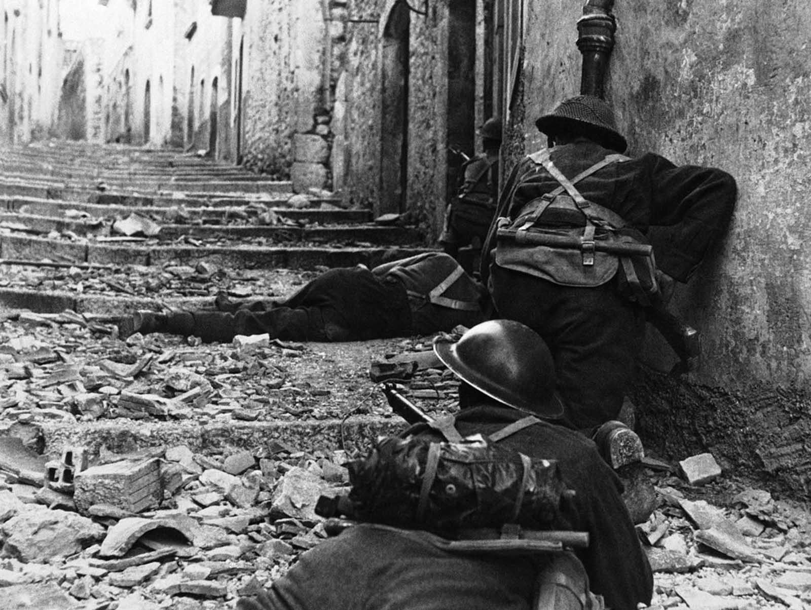 Over the body of a dead comrade, Canadian infantrymen advance cautiously up a narrow lane in Campochiaro, Italy, on November 11, 1943. The Germans left the town as the Canadians advanced, leaving only nests of snipers to delay the progress.