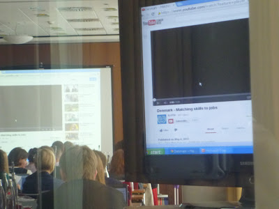 Angeschnitten ein Flachbildschirm, im Hintergrund Konferenzbesucher