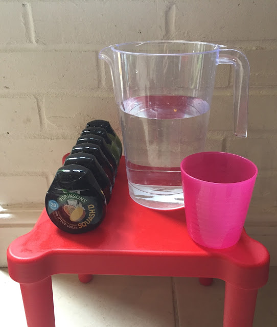 A small red table with 6 Robinsons squash'd bottles, a clear plastic jug of water and a pink cup