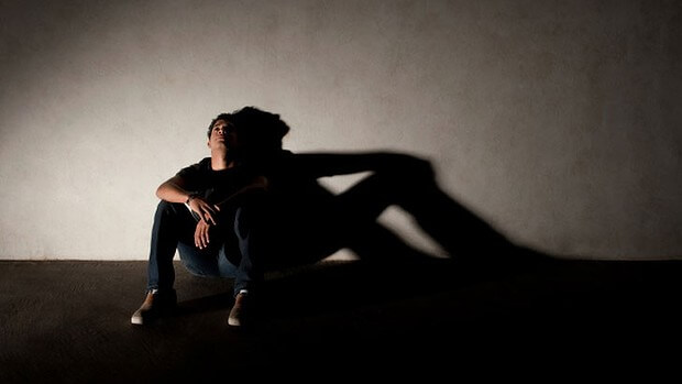 Hombre joven sentado en el suelo, solo, con su sombra a la derecha de la imagen.