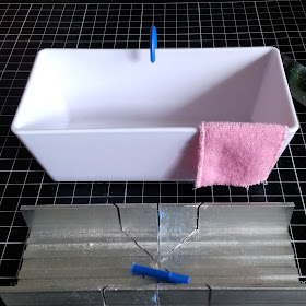 One-twelfth scale modern miniature bath with pink towel over the side and blue tap over the back, sitting on a cutting mat with a miniature mitre box in front of it.