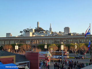 Coit Tower pierces the blue sky to the left of the Transamerica building. Buses, cars and many people are visible in the foreground.