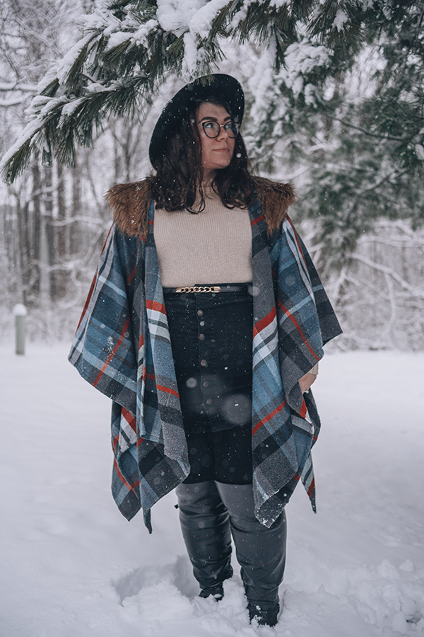 An outfit of a black wide brim hat, gray, red, and blue plaid shawl with brown faux fur collar, cream sweater, black button up mini skirt, and black over the knee boots in snow.