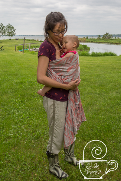 [Image of a tan skin bespectacled Asian woman smiling down at the toddler on her hip wrapped in a coral and silver painterly pattered woven wrap carrier. The woman is wearing a bird-print maroon shirt and beige-ish tencel pants with black and white rain boots. They're standing on grass in front of a lake on a cloudy afternoon.]