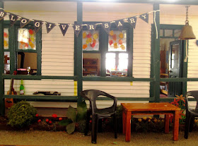 One-twelfth scale miniature school house exterior with a bunting strung across the verandha spelling out 'blogiversary'. Chairs and a table are set up outside the school, while on the veranda is a guitar, a ukulele, a bottle fo wine and a wine glass.