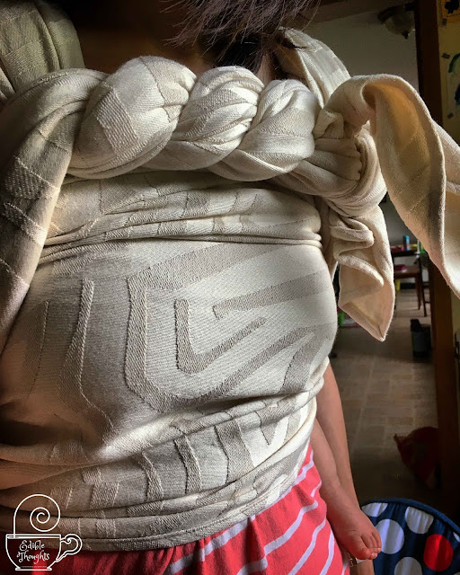[Image of a woman's torso with an ecru woven wrap baby carrier spread across the chest to show off the wrap's abstract geometric pattern in cloudy lighting. The double hammock back carry is finished with a twist across the top called a candy cane chest belt. It looks and feels so luxurious but without the fussy care of what I would think something luxurious would need.]