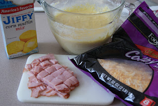 Corn bread mix in a clear mixing bowl, with a bag of shredded cheese, along side chopped ham on a cutting board.