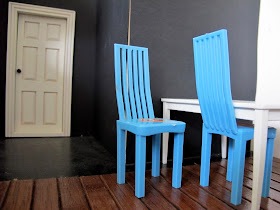 Two blue plastic modern miniature dolls house chairs, at a white table against a black wall and wooden floor.