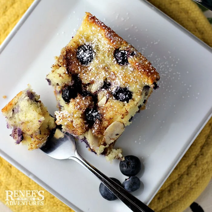 Overhead view of blueberry coffee cake piece on white plate with a bite ready to eat sitting on tines of fork