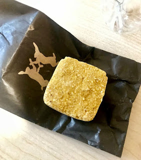 A bright gold square compact scrub full of little whiite pieces of sugar and oatmeal on top of a rectangular black bag on a white rectangular table on a bright background 