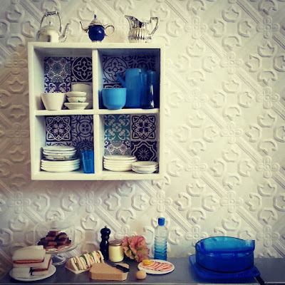 Modern miniature white wall shelf with blue and white tiles on the back, filled with blue and white crockery and with various jugs displayed on the top. Underneath, on a metal bench top, are the plates of cakes and sandwiches, with the sandwich components laid out. 