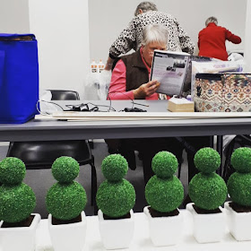 Six one-twelfth scale topiaries in planters lined up on a table. In the background is a woman reading a magazine and two women with their backs turned to the camera working on something.