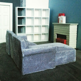 One-twelfth scale modern miniature lounge scene with a grey velvet sectional sofa in front of a fireplace.