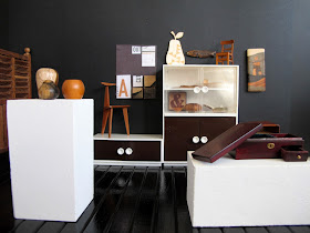 Modern dolls' house miniature gallery, showing various wooden furniture and homeware pieces displayed on plinths, in a glass-fronted  cupboard, and on the wall.