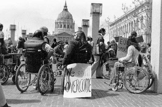 A diverse crowd of people with disabilities gathers in front of the HEW building, city hall can be seen in the distance, a sign reads "We Shall Overcome."