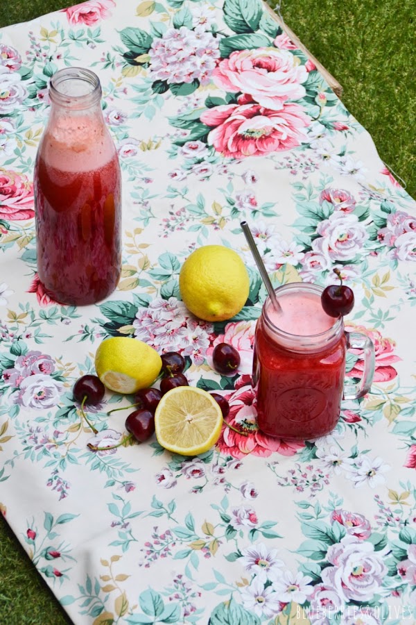MANTEL DE FLORES SOBRE CÉSPED, BOTELLA E CRISTAL LLENA DE LIMONADA DE CEREZAS Y JARRA DE CRISTAL, CEREZAS Y RODAJAS LIMÓN