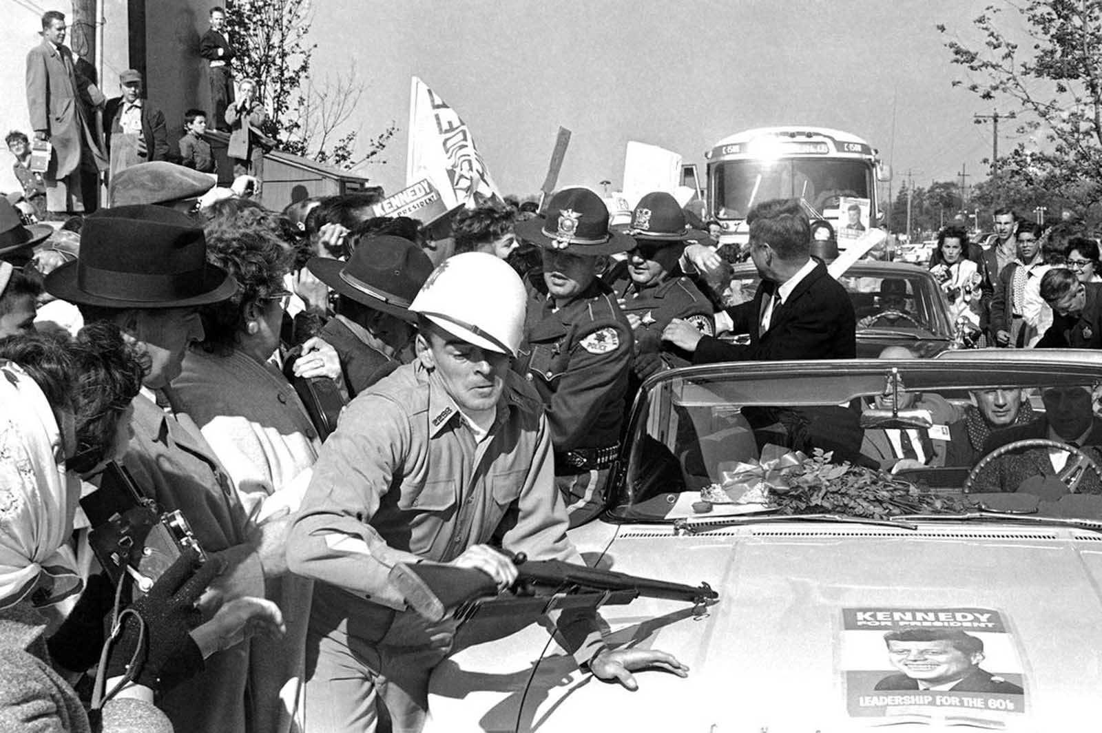 Helmet askew, rifle definitely not at the ready, man in military uniform is jammed against fender of car bearing the Democrats' presidential nominee, on October 25, 1960 in Elgin, Illinois. 