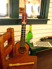 A miniature dolls' house veranda, with seating, a bottle and glass of wine, a recorder, guitar, ukulele and sheet of music.