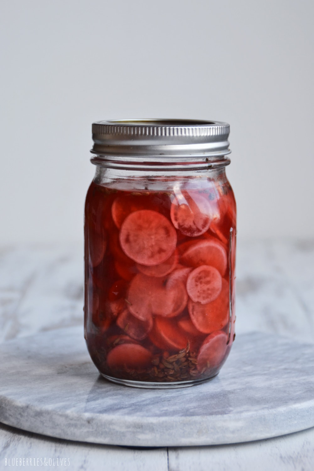 PICKLED RADISHES IN A GLASS JAR WITH LID, WHITE MARBLE CUTTING BOARD AND WHITE BACKGROUND