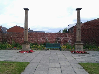 Two pillars in a walled garden, a bench between.  The bases of the pillars are inscribed with names and there are poppy wreaths around the bottoms of the pillars.