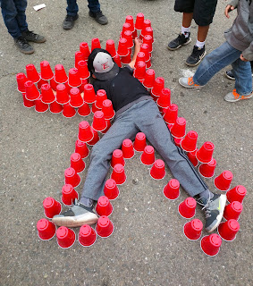 Boy lying on concrete inside body outline made of red dixie cups, other boys looking but only their feet are visible