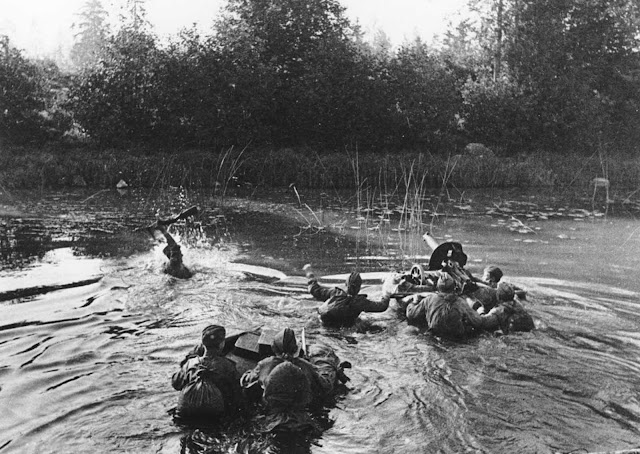A Soviet machine gun crew crosses a river along the second Baltic front, in January of 1945. The soldier on the left is holding his rifle overhead while his comrades push a floating device with the a Maxim machine gun, followed by two men with several supply boxes.