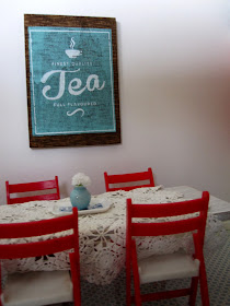 Modern one-twelfth scale miniature dining table with a cream lace cloth and red fold-up chairs set around it.