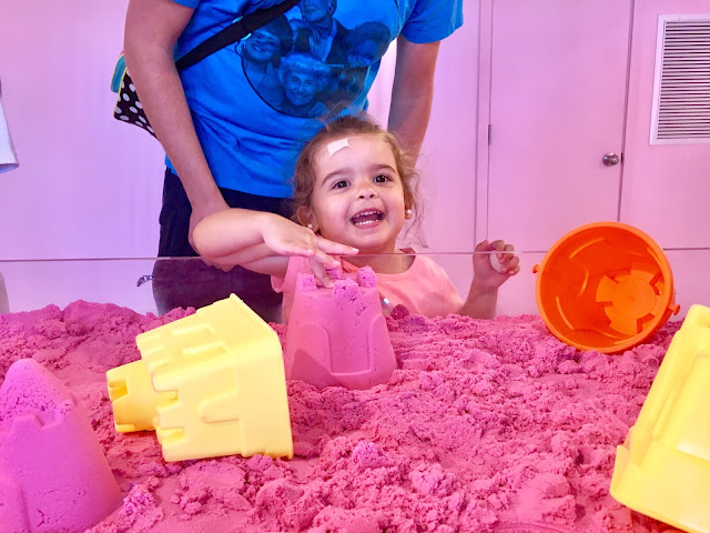 Toddler girl smiles and points to a pink kinetic sand castle that she built