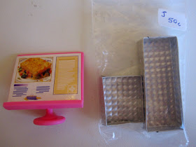 Pink plastic miniature menu board and bag containing two silver-coloured miniature trays. Bag is marked 'J 50c'