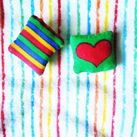 Two one-twelfth scale cushions, one with a rainbow print, the other with a heart print, on a rainbow-striped background.