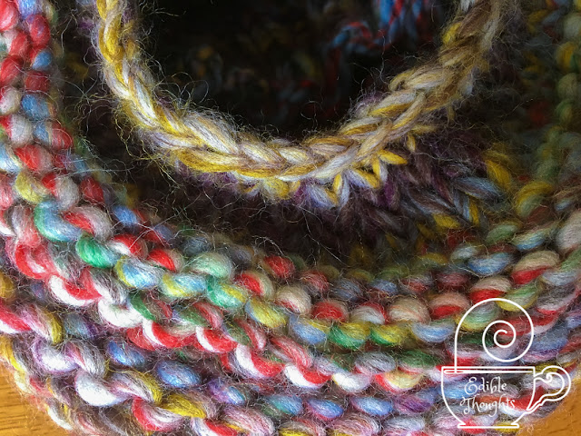 [Image is a close-up of a multi-colored chunky wool yarn knit hat alternating bands of purl and knit rounds with a hole on top. Bottom right corner watermark in white is a tea cup with a needle lying horizontally across the rim with a swirl of thread coming off the top like steam. In white text on the cup is Edible Thoughts copyright symbol Stephanie Lu Sinclair.]