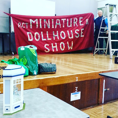 Man putting up a fabric banner for the ACT Miniature & Dollhouse Show on a stage in a community hall. Boxes and bags and unpacked miniatures lie on the stage and table in front of it.