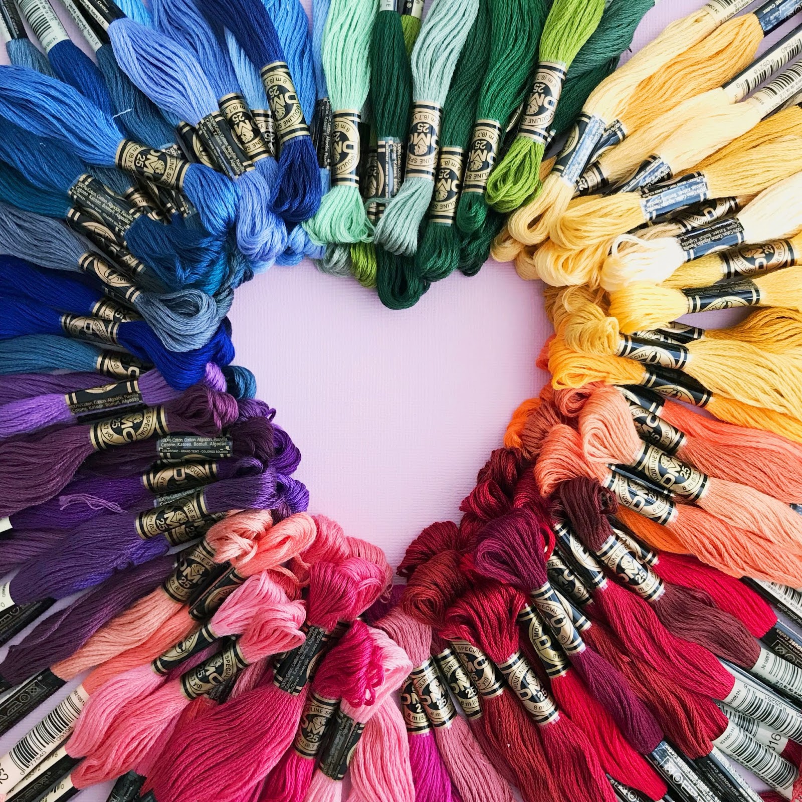 A photo of lots of DMC floss spread around the outside of the photo, in rainbow colour order, with the space in the middle in the shape of a heart.