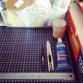 Cutting mat with a ruler, tweezers, cutting knife and tube of Weldbond glue arranged on it. To the right is a package of baking paper and to the back is a tub of miniature kits.