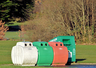 recycling bins sitting at the edge of pavement with grass and trees in background.