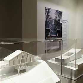 Architect's models of two modernist building in perspex cases in front of a large photo and information about one of them on the wall behind.