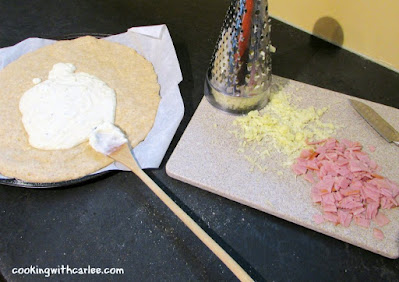 croque madame pizza being assembled beschamel spread over pizza crust with shredded cheese and ham nearby