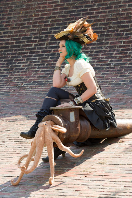 Woman dressed as a Steampunk Pirate with green hair, green lipstick, pirate's tricorn hat, hi lo hem skirt, blouse, corset, boots and pet stuffed animal squid/octopus