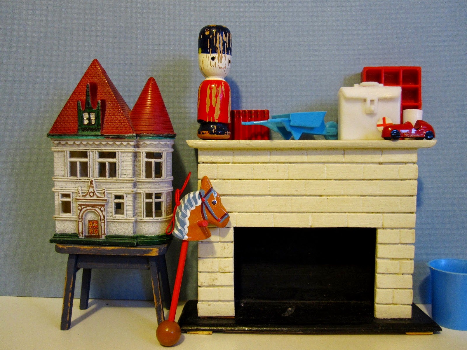 Selection of vintage miniature accessories displayed around a dolls' house mantlepiece, with a dusky-blue coloured wall behind.