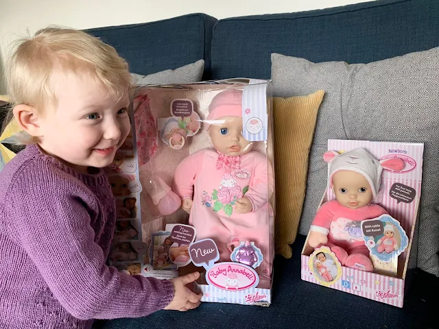 A toddler next to Baby Annabell and Baby Annabell Sweetie in their packaging ready for review