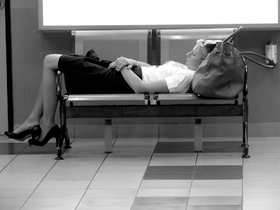Eine am Flughafen hängengebliebene Dame liegt auf einer Sitzbank, die Reisetasche dient als Kopfkissen
