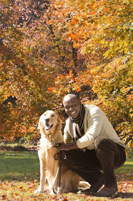 A Black man crouches next to his dog, a Golden Retriever,  in the park in Autumn