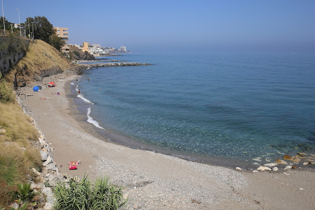 Playa de piedras y arena gruesa, con acantilados a su espalda y las aguas cristalinas del mar a su frente.