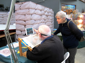 A woman watching a man stitching a very fine miniature carpet.