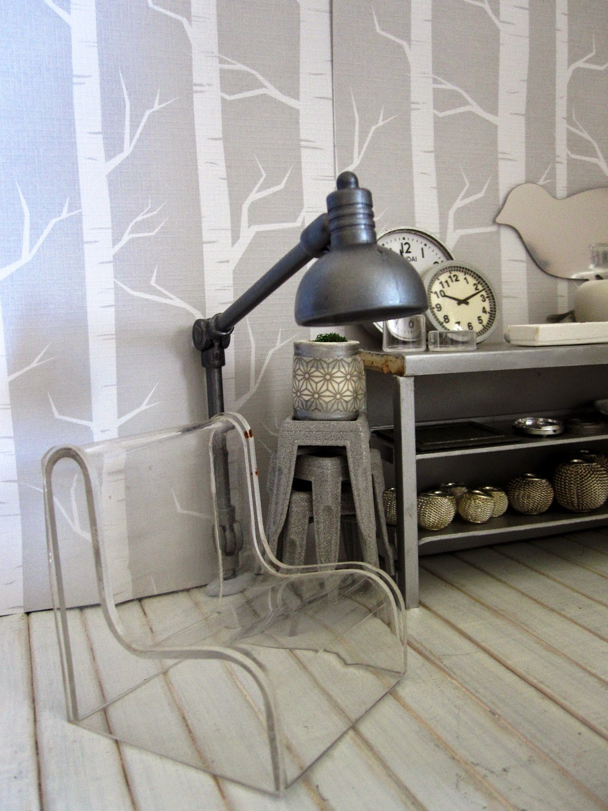 Corner of a modern dolls' house miniature homeware shop in grey and white. On display is a clear perspex chair, a giant floor lamp, a stack of cafe stools and a grey metal shelving unit with various grey-coloured decor pieces displayed.