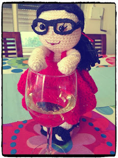 Kwokkie Doll is standing on a table next to a glass of white wine.