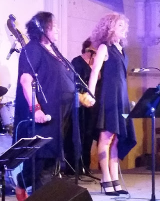 Vonda Last (left) and Sandra Pires (right) are standing on stage facing the audience holding hands and smiling to audience applause